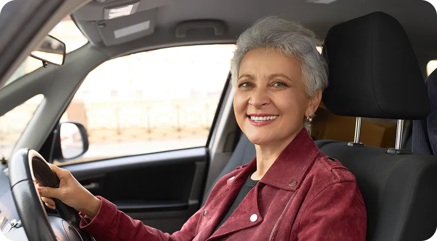 A woman with short gray hair smiles while sitting in the driver's seat of a car.