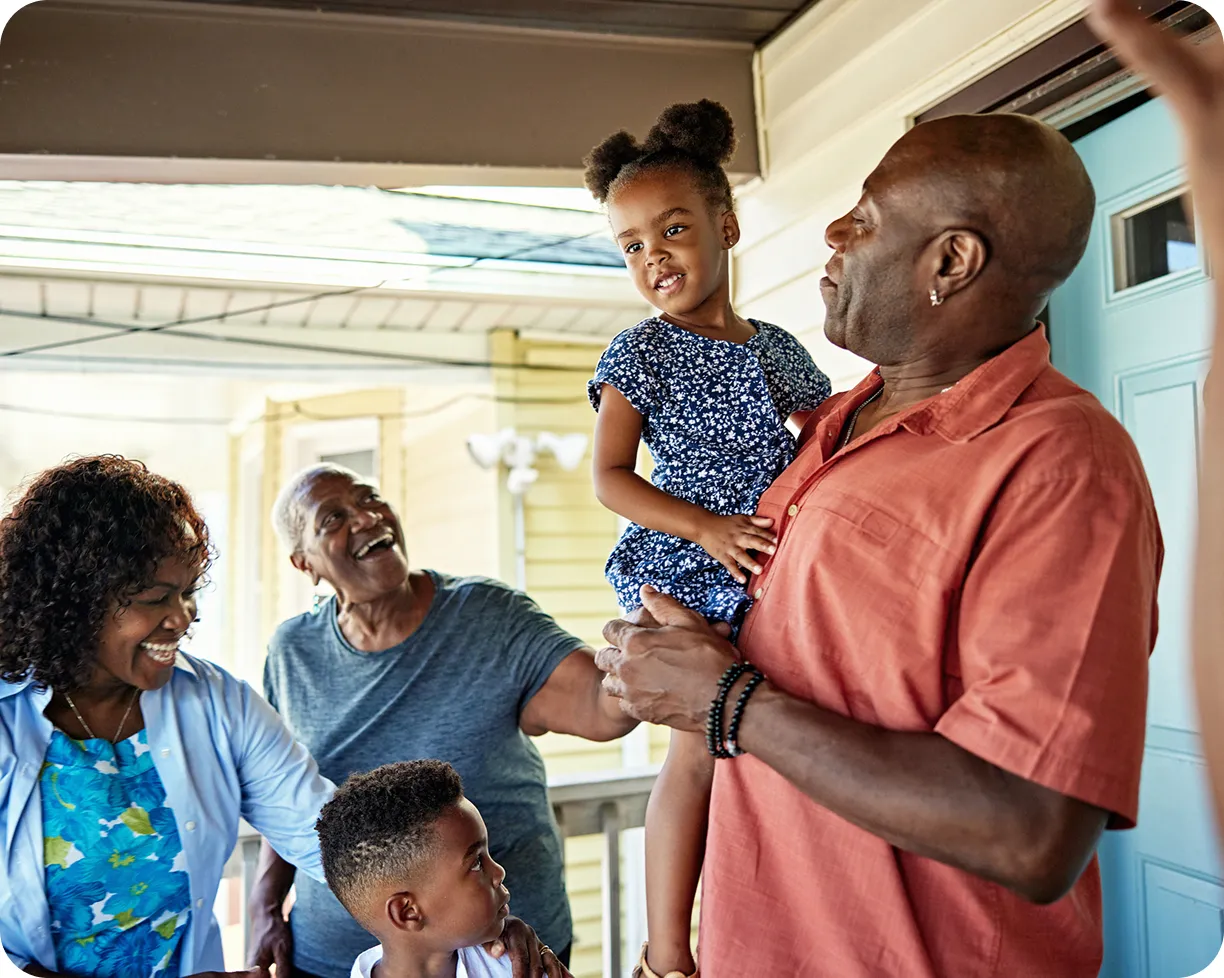 A man holds a smiling young girl while family members gather around on a porch.