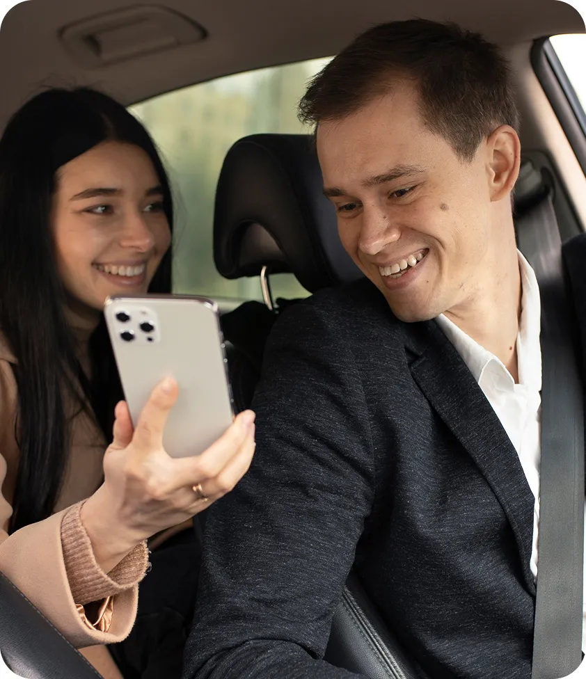 Smiling couple sitting in a car, sharing something on a phone while wearing seat belts.