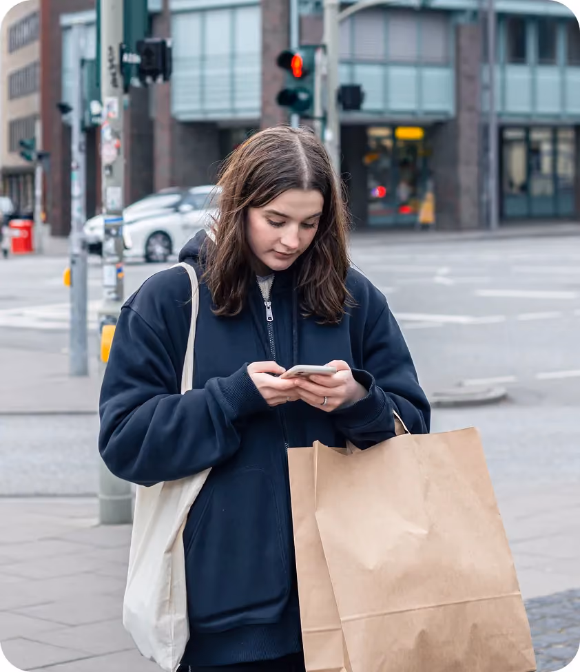 Young woman walking in the city with shopping bags, looking at her phone and wearing a blue hoodie.