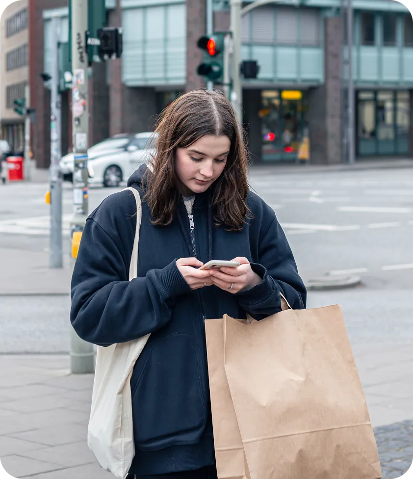 Young woman walking in the city with shopping bags, looking at her phone and wearing a blue hoodie.
