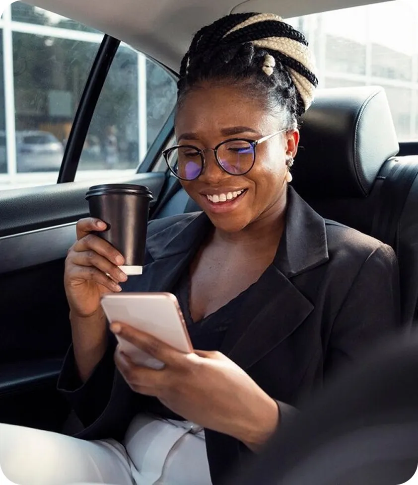 Businesswoman sitting in the back seat of a car, smiling at her phone while holding a coffee cup.