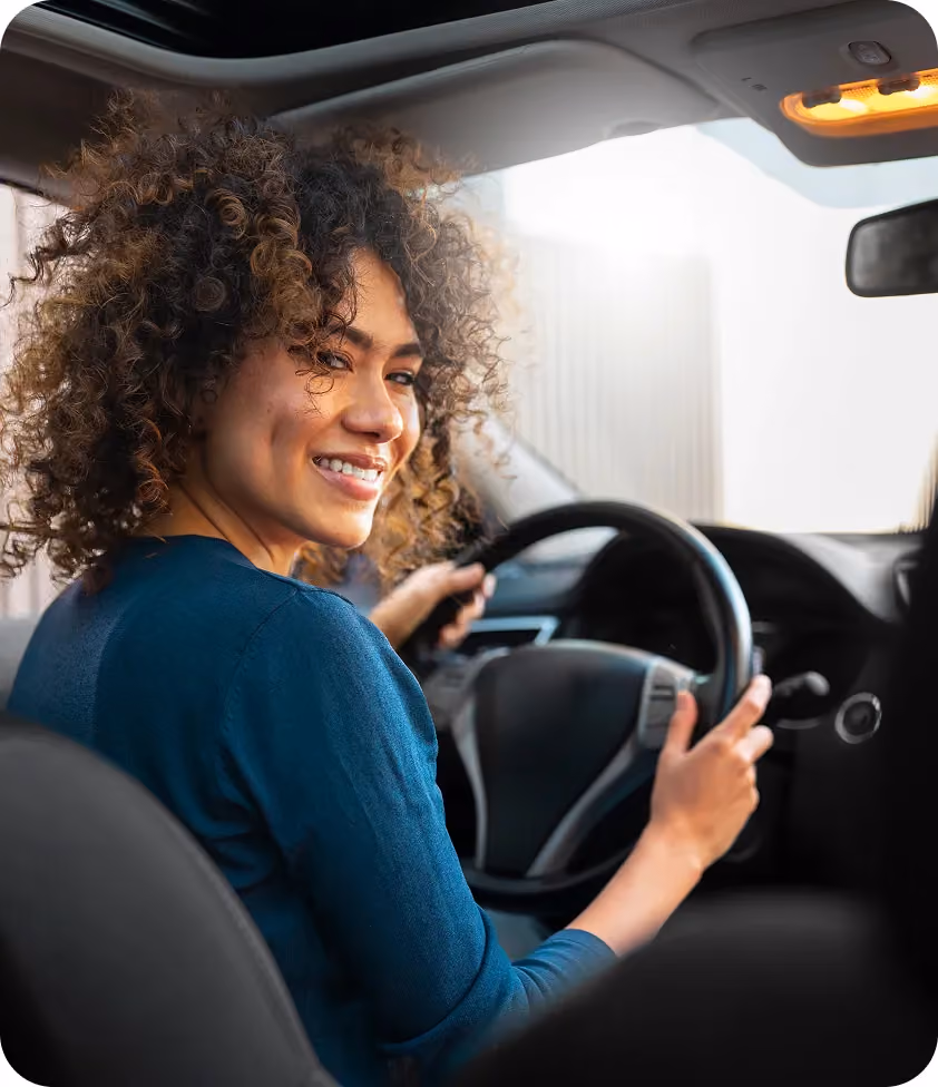 Smiling woman with curly hair sitting in the driver’s seat of a car, looking over her shoulder.