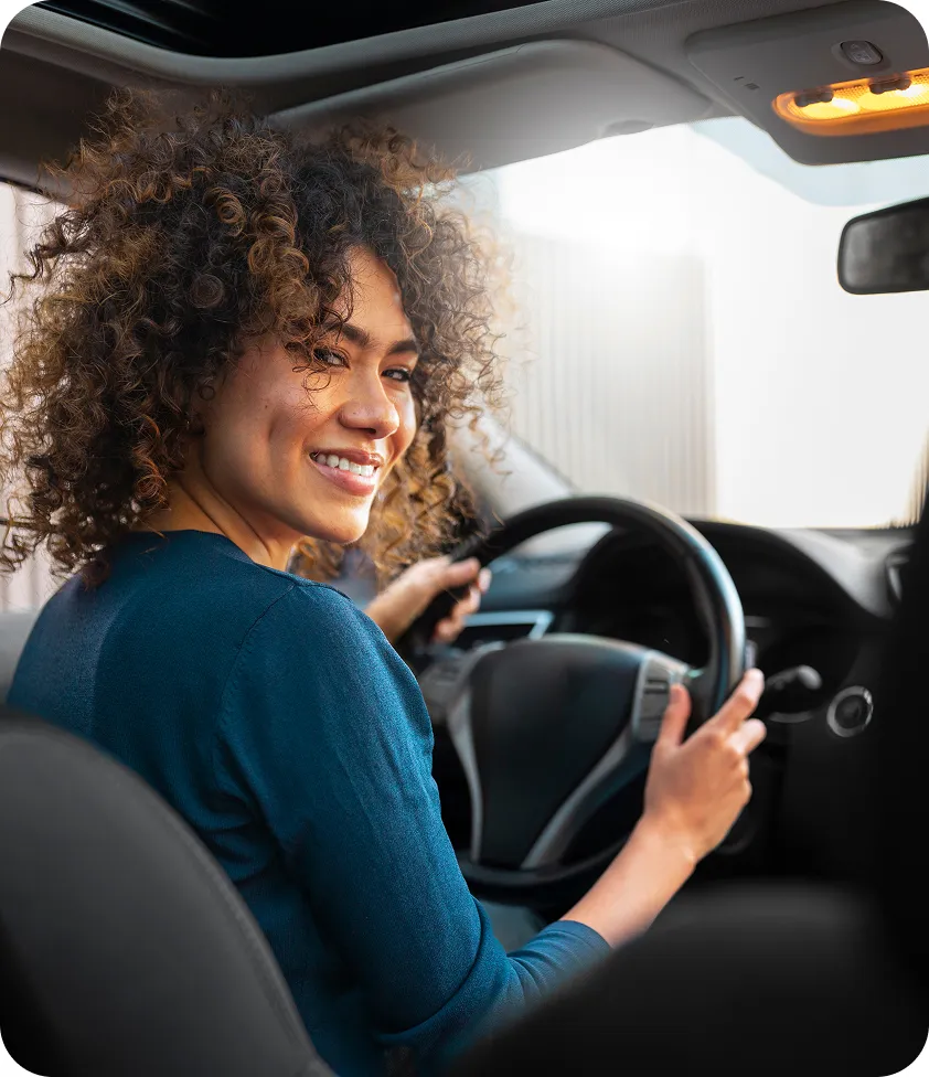 Smiling woman with curly hair sitting in the driver’s seat of a car, looking over her shoulder.