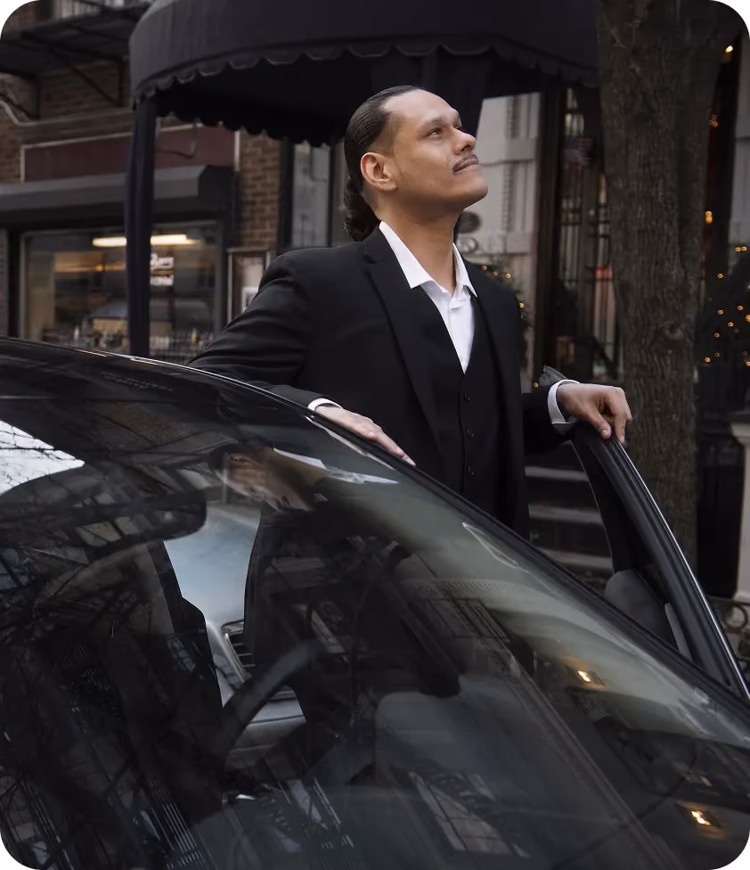 Man in a suit standing beside a car on a city street, looking up with a confident expression
