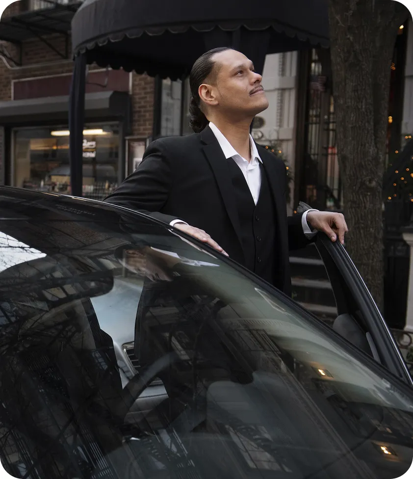 Man in a suit standing beside a car on a city street, looking up with a confident expression