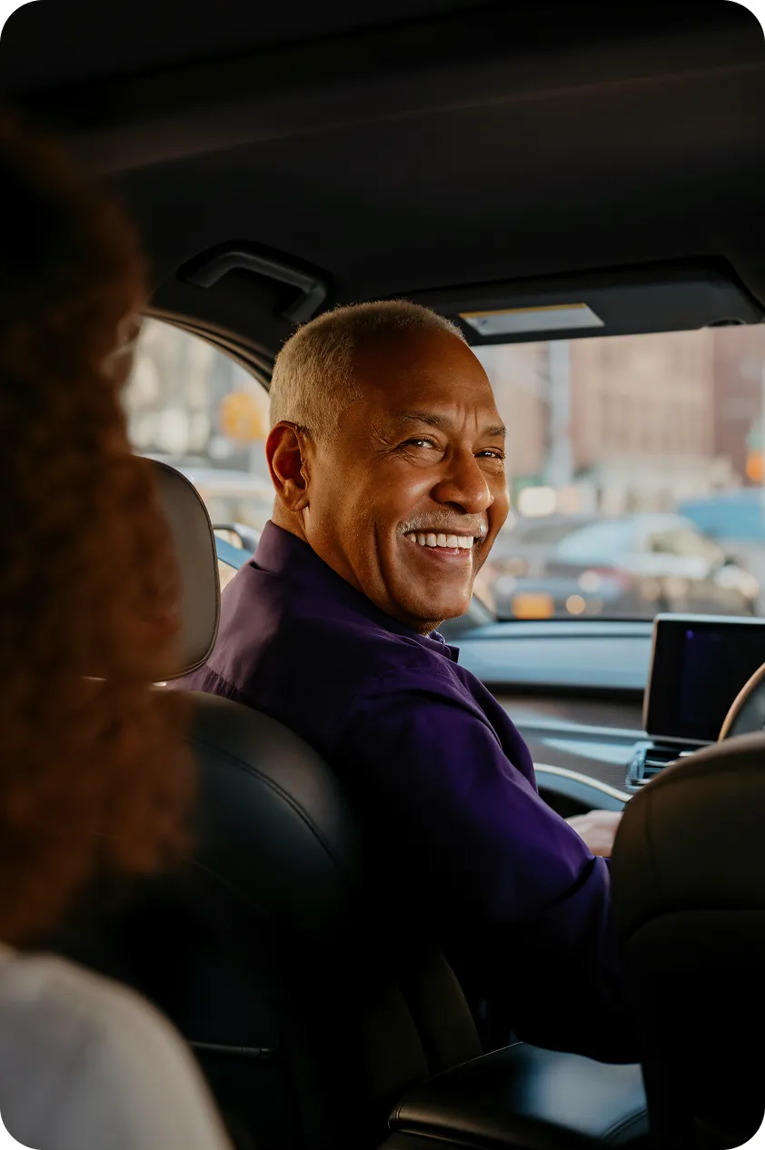 A man in a purple shirt smiles while driving a car, looking at the passenger.