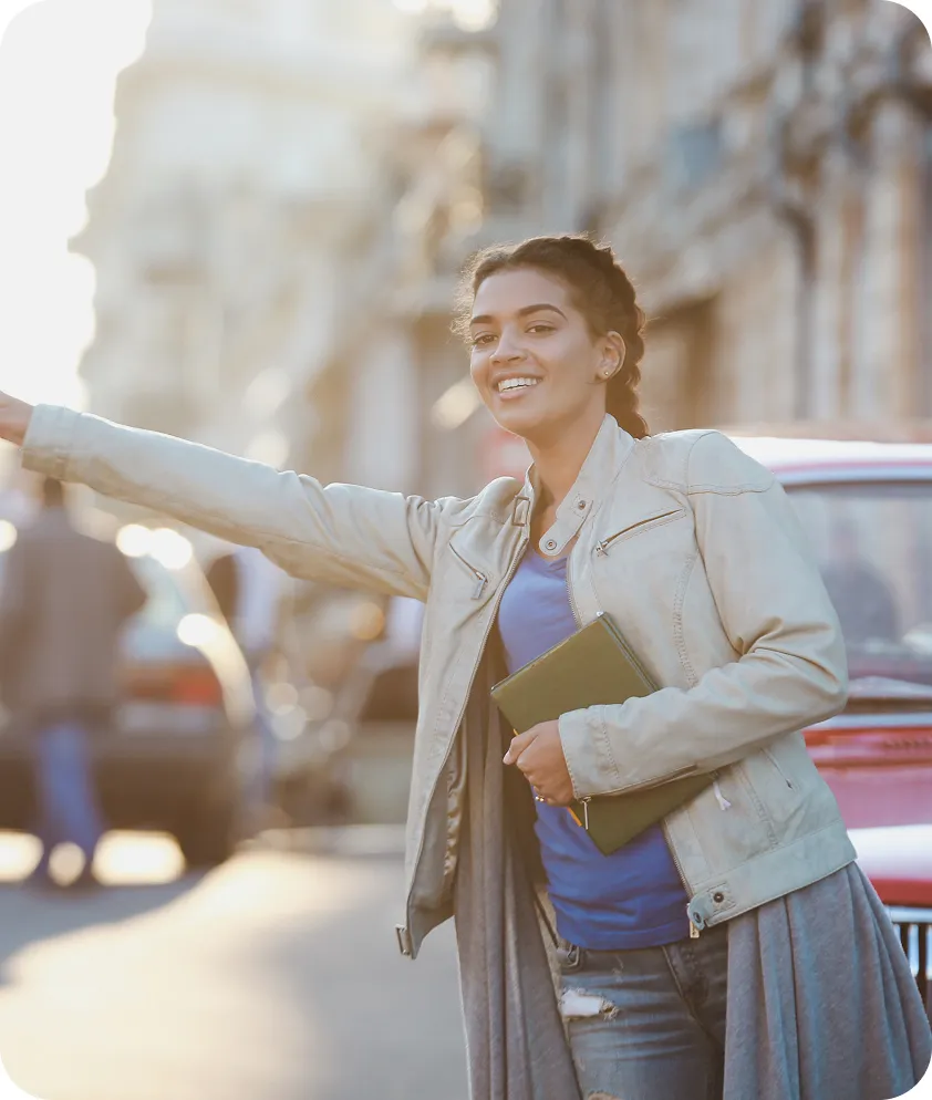 A person stands on the street, smiling and hailing a taxi, holding a notebook.