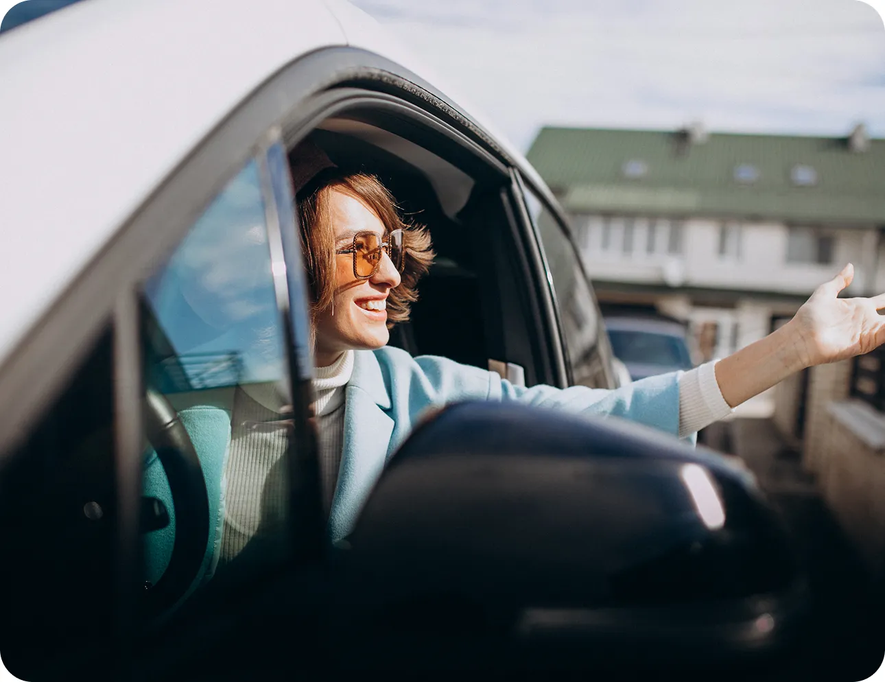 Person wearing sunglasses leans out car window, smiling and gesturing with one hand.