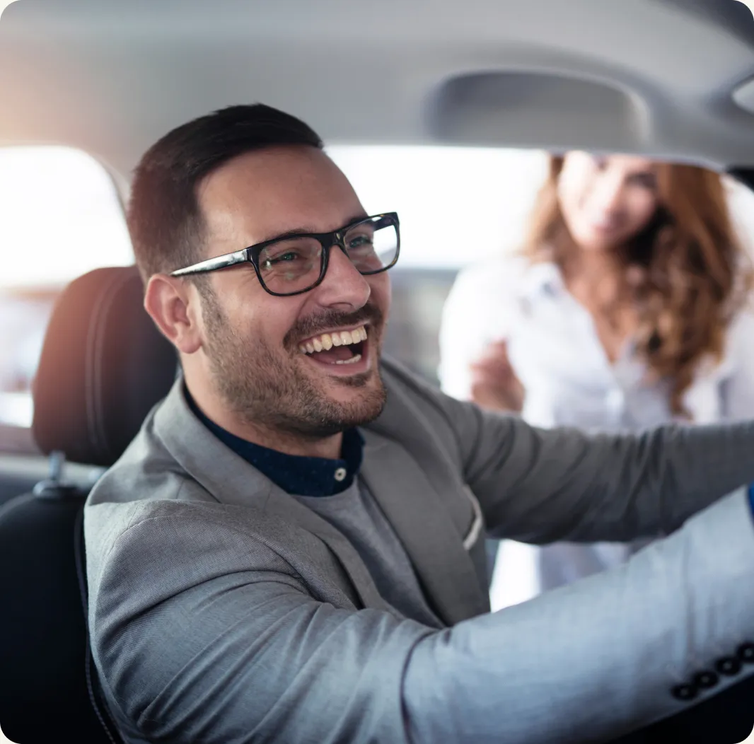 A man in glasses smiles while sitting in the driver's seat of a car.