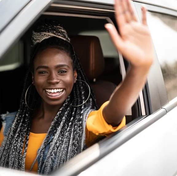 A woman with braided hair is smiling while waving from a car window.