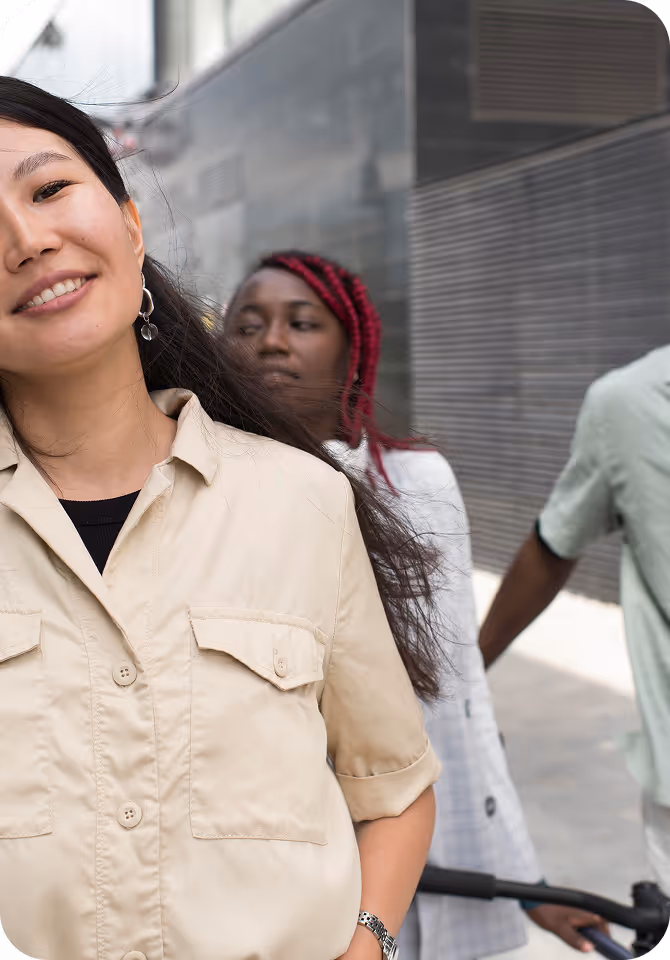 Three people smiling and walking on an urban street; a woman in a beige shirt is in the foreground, while two others are behind her.