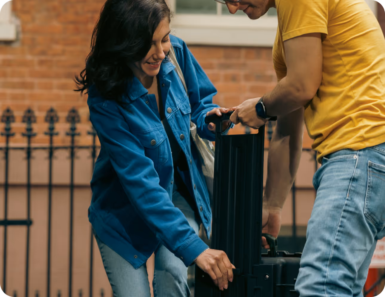 Two people outdoors handling a suitcase; a woman in a blue jacket smiles while helping, and a man in a yellow shirt assists.