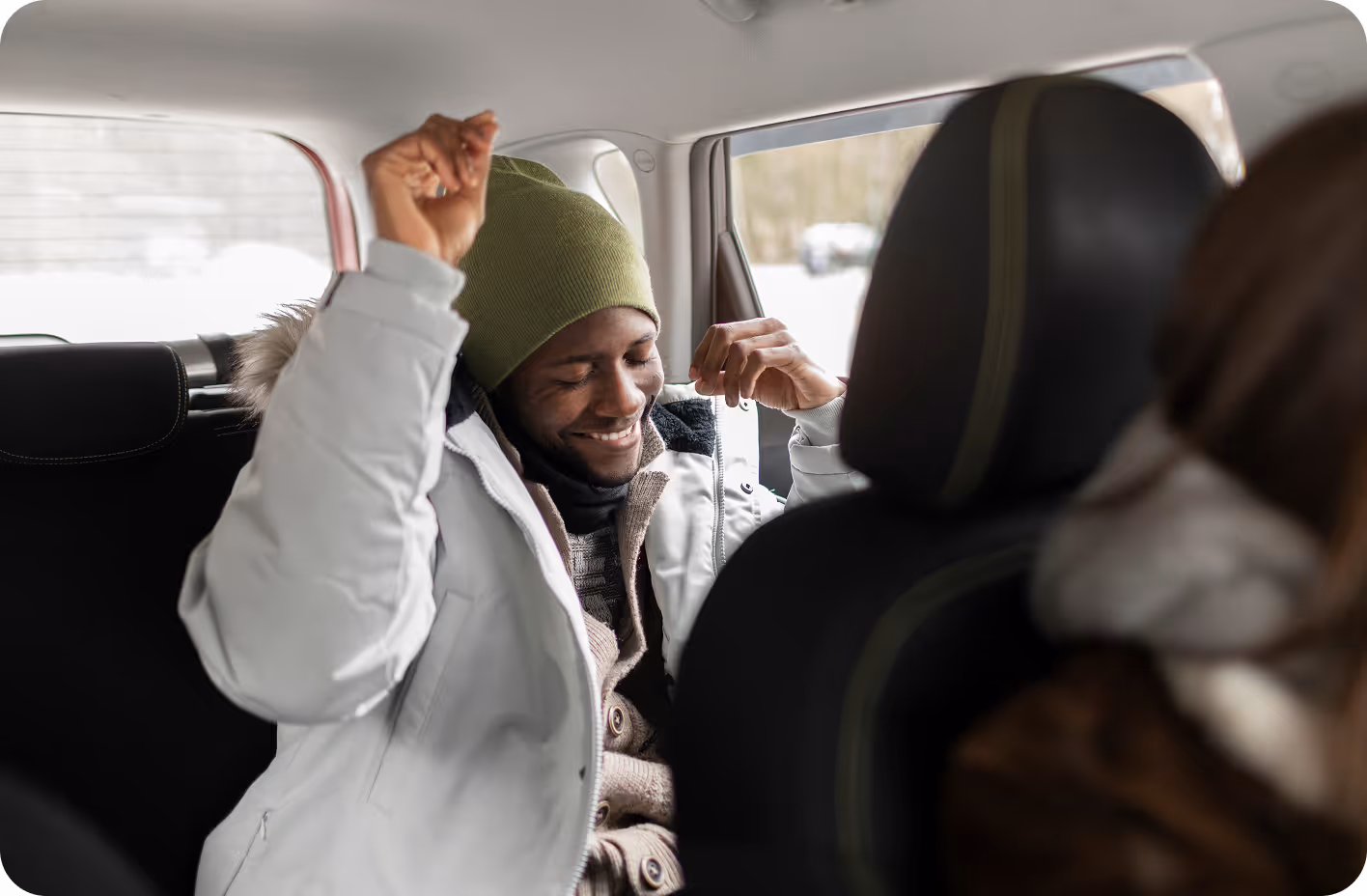 A man wearing a green beanie and a gray winter jacket is sitting in the backseat of a car, smiling with his eyes closed and raising his arms as if dancing or celebrating.