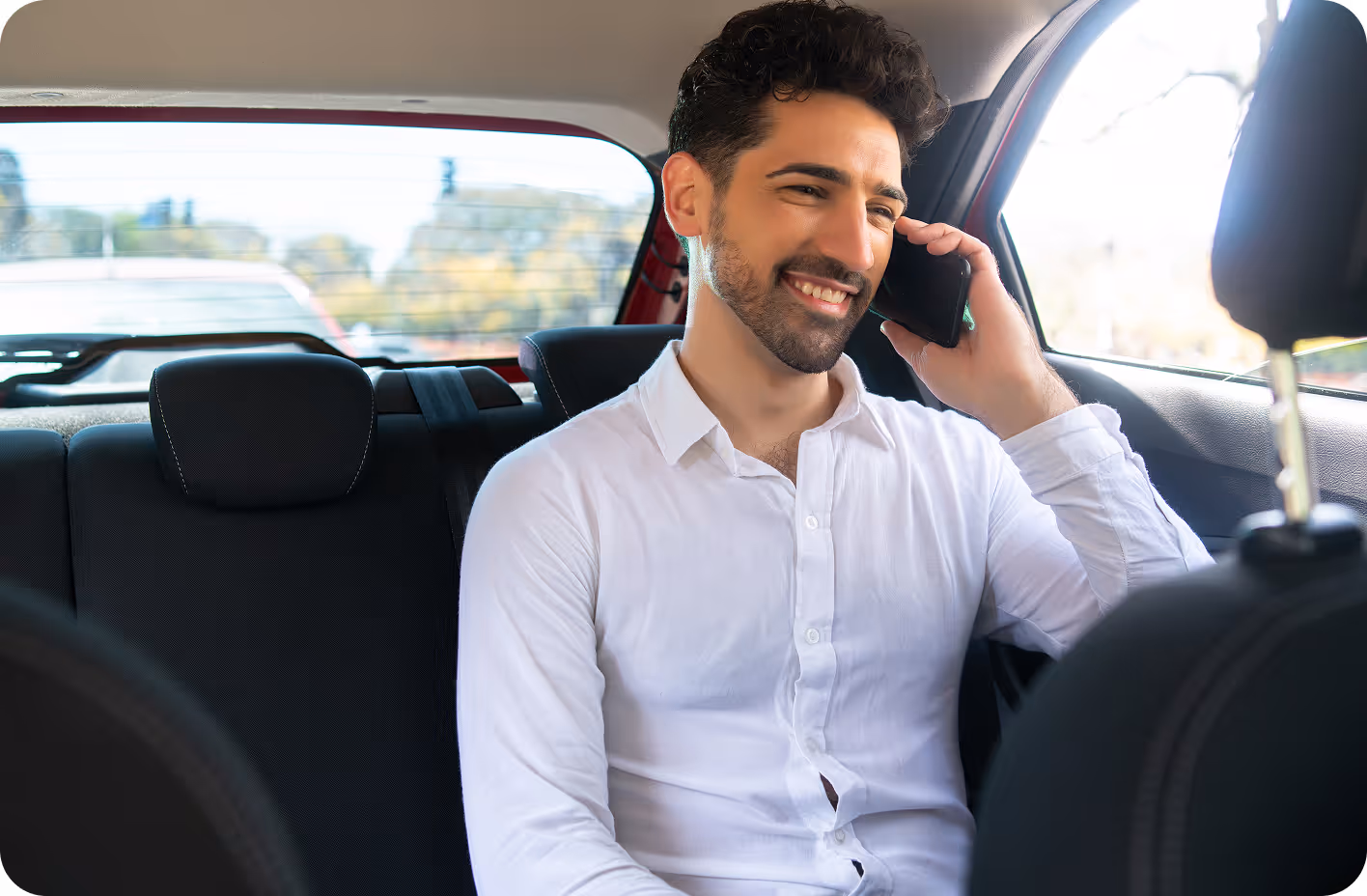 A man in a white shirt smiles while talking on his cellphone, sitting in the backseat of a car.