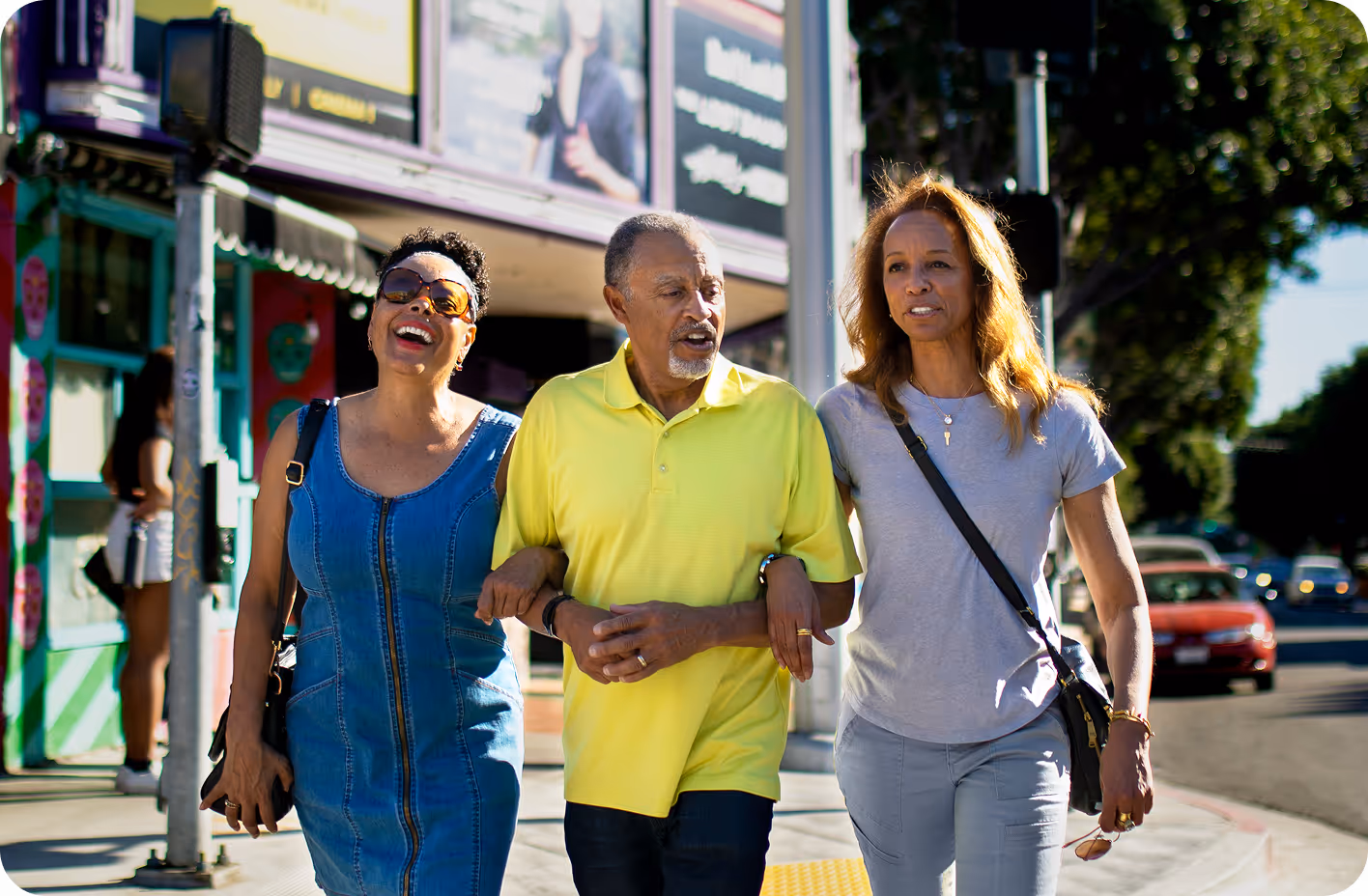 An elderly man in a yellow polo shirt walks arm in arm with two women, all smiling, as they cross a sunny urban street with colorful surroundings.