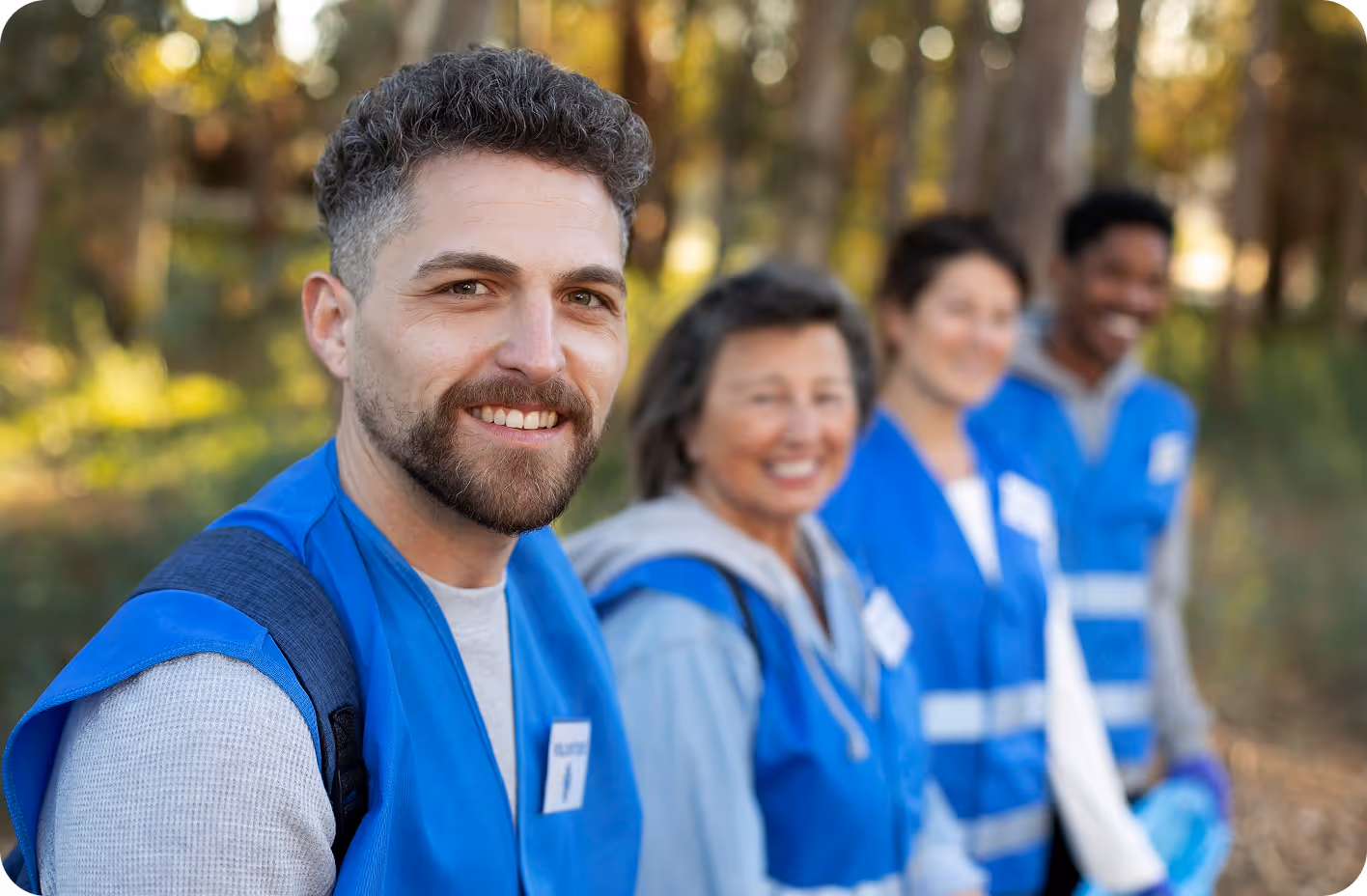 A smiling man wearing a blue volunteer vest stands outdoors with three other volunteers blurred in the background, all dressed similarly, suggesting teamwork and community service.