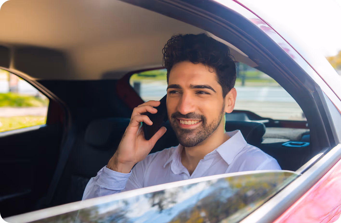 A man in a white shirt smiles while talking on his cellphone, sitting in the backseat of a car.