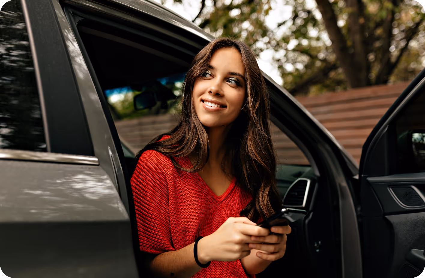 A young woman in a red sweater sits at the open door of a car, holding a smartphone and smiling while looking off to the side.