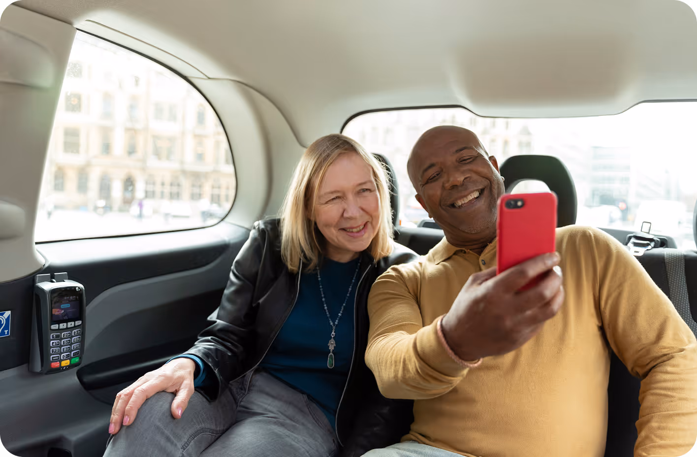 A smiling man holds a red smartphone to take a selfie with a woman sitting beside him in the backseat of a car, both appearing happy and relaxed