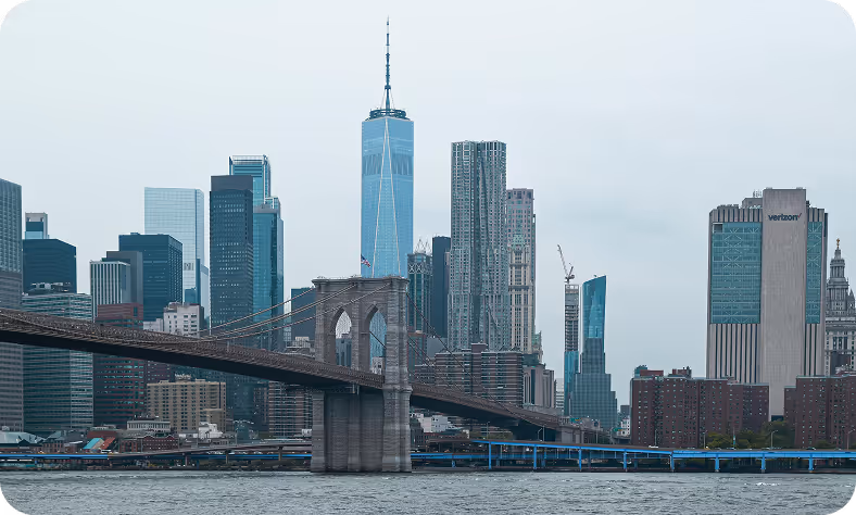 The Brooklyn Bridge stretches across the East River with the New York City skyline in the background, featuring the One World Trade Center and other tall skyscrapers.