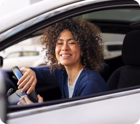 A smiling woman with curly hair sits in the driver’s seat of a car, holding the steering wheel and looking out of the open window.