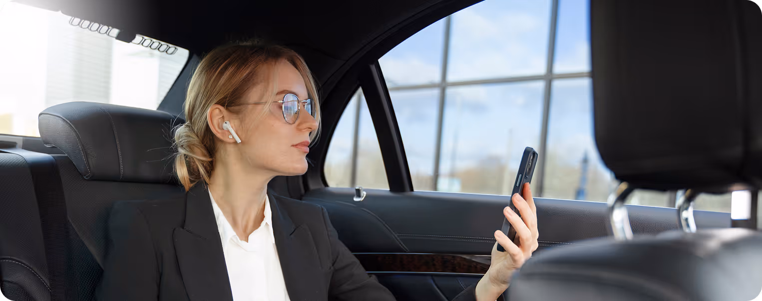 A woman in glasses and a black blazer sits in the backseat of a car, holding her smartphone and wearing wireless earbuds, appearing focused.