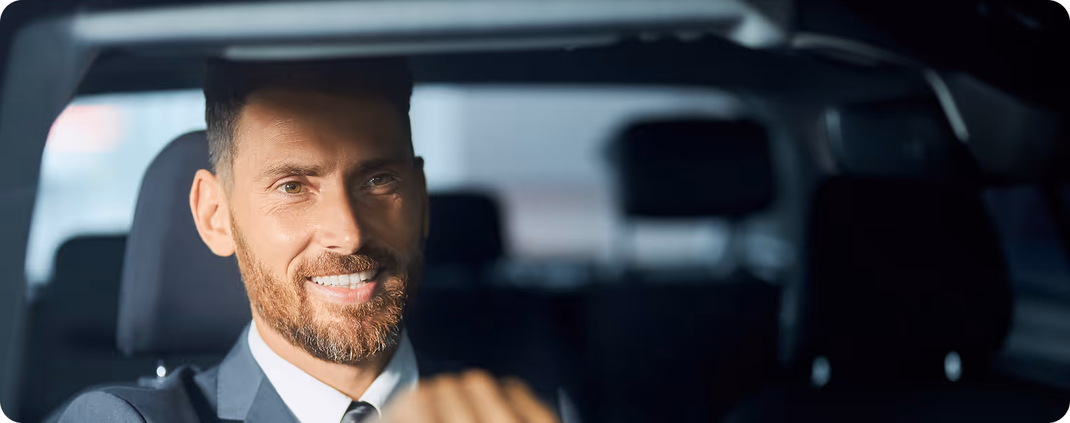 A man in a suit sits in the driver’s seat, smiling confidently as he looks ahead while driving.
