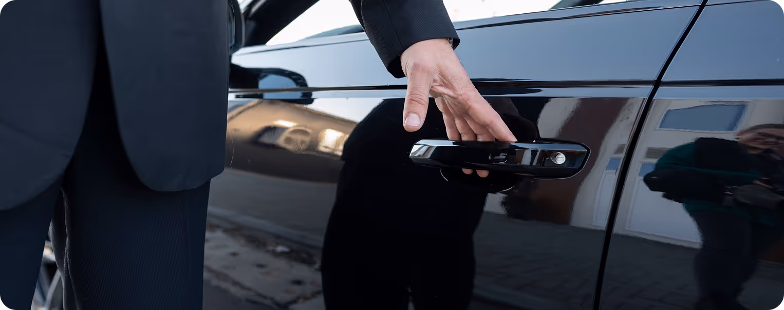A person in a suit reaches to open the handle of a black car door, with the reflection of a photographer visible on the vehicle.