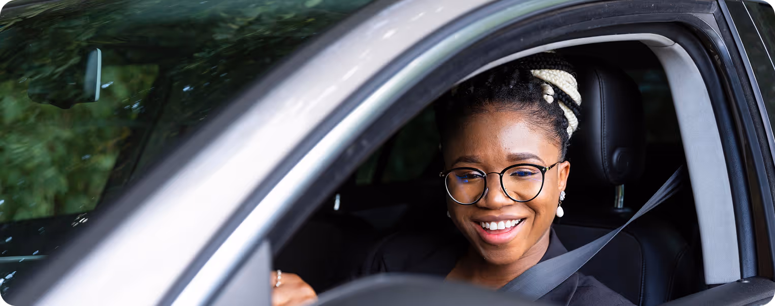 A woman wearing glasses and a black blazer smiles while sitting in the driver’s seat of a car, with her seatbelt fastened.