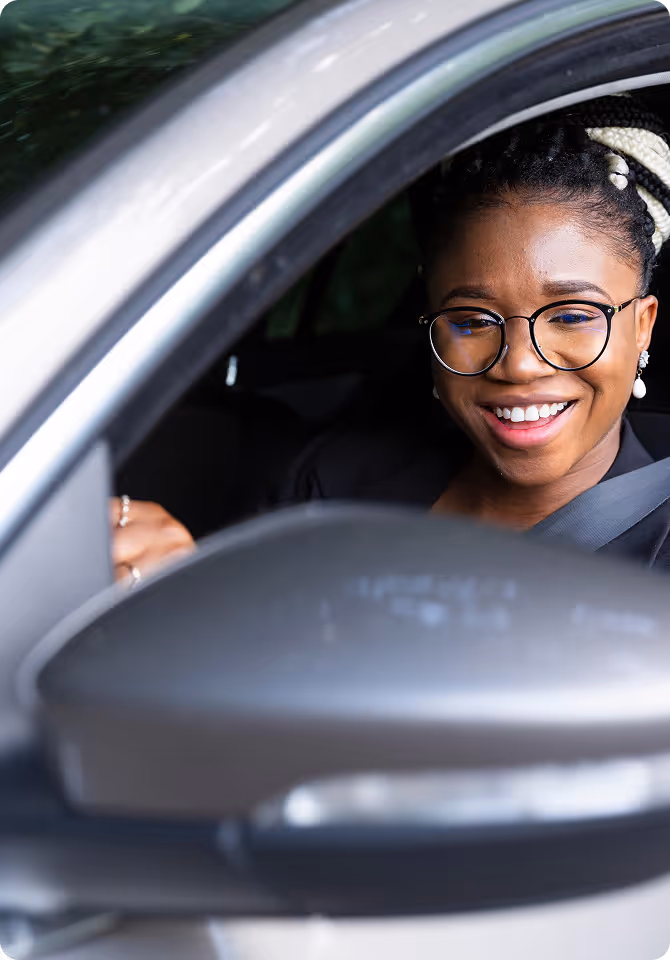 A woman wearing glasses and a black blazer smiles while sitting in the driver’s seat of a car, with her seatbelt fastened.