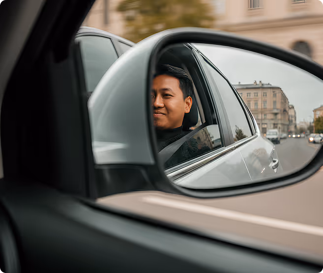 Side mirror reflection showing a man in a car wearing a seatbelt with a city street in the background.