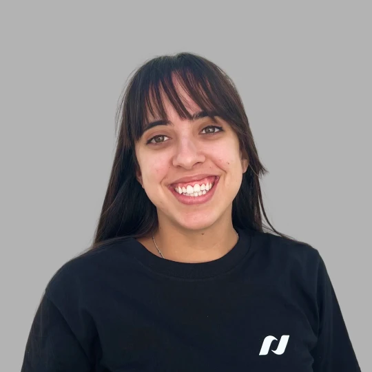 Smiling young woman with long dark hair and bangs wearing a black shirt with a small white logo on a gray background.