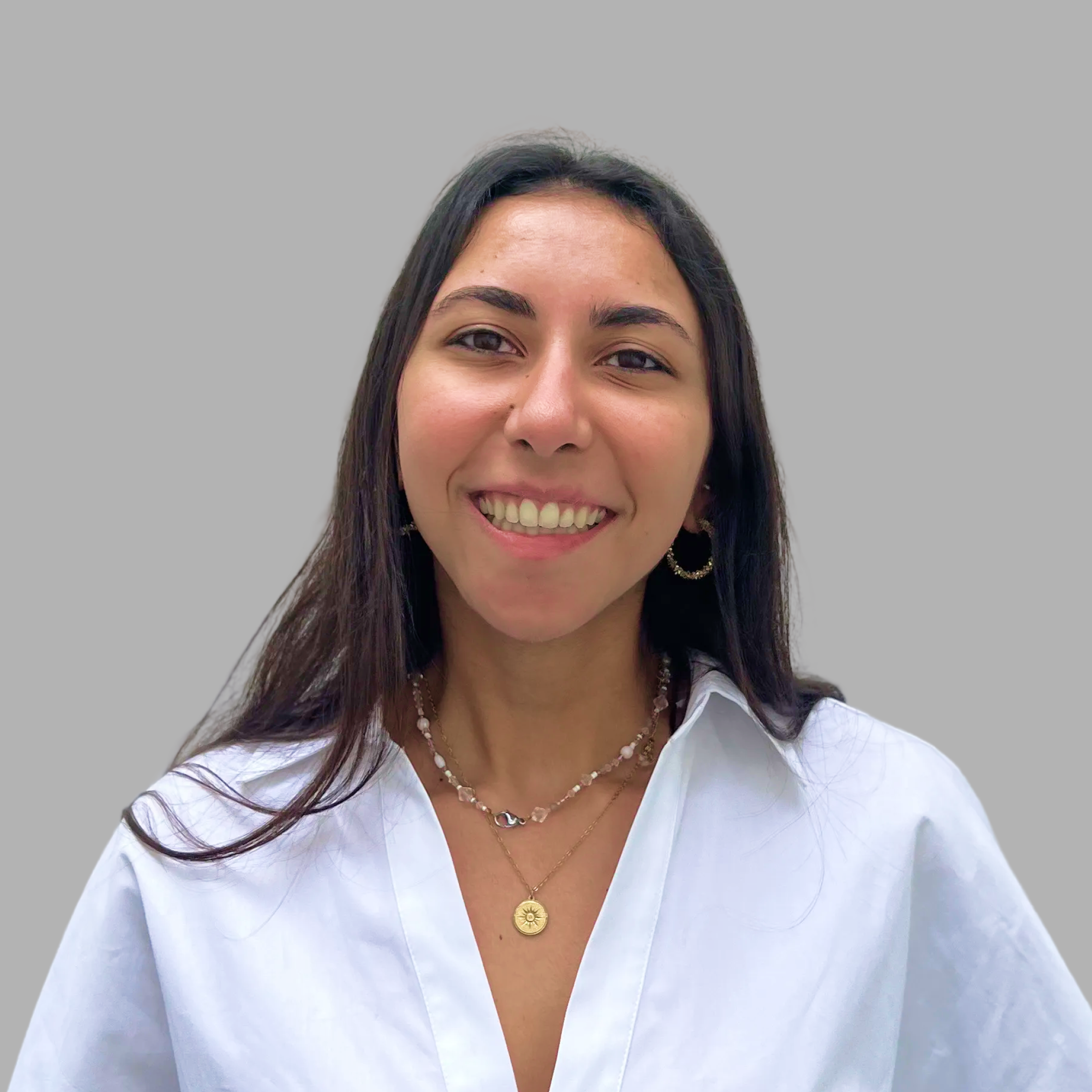 Smiling woman with long dark hair wearing a white shirt and layered necklaces against a gray background.