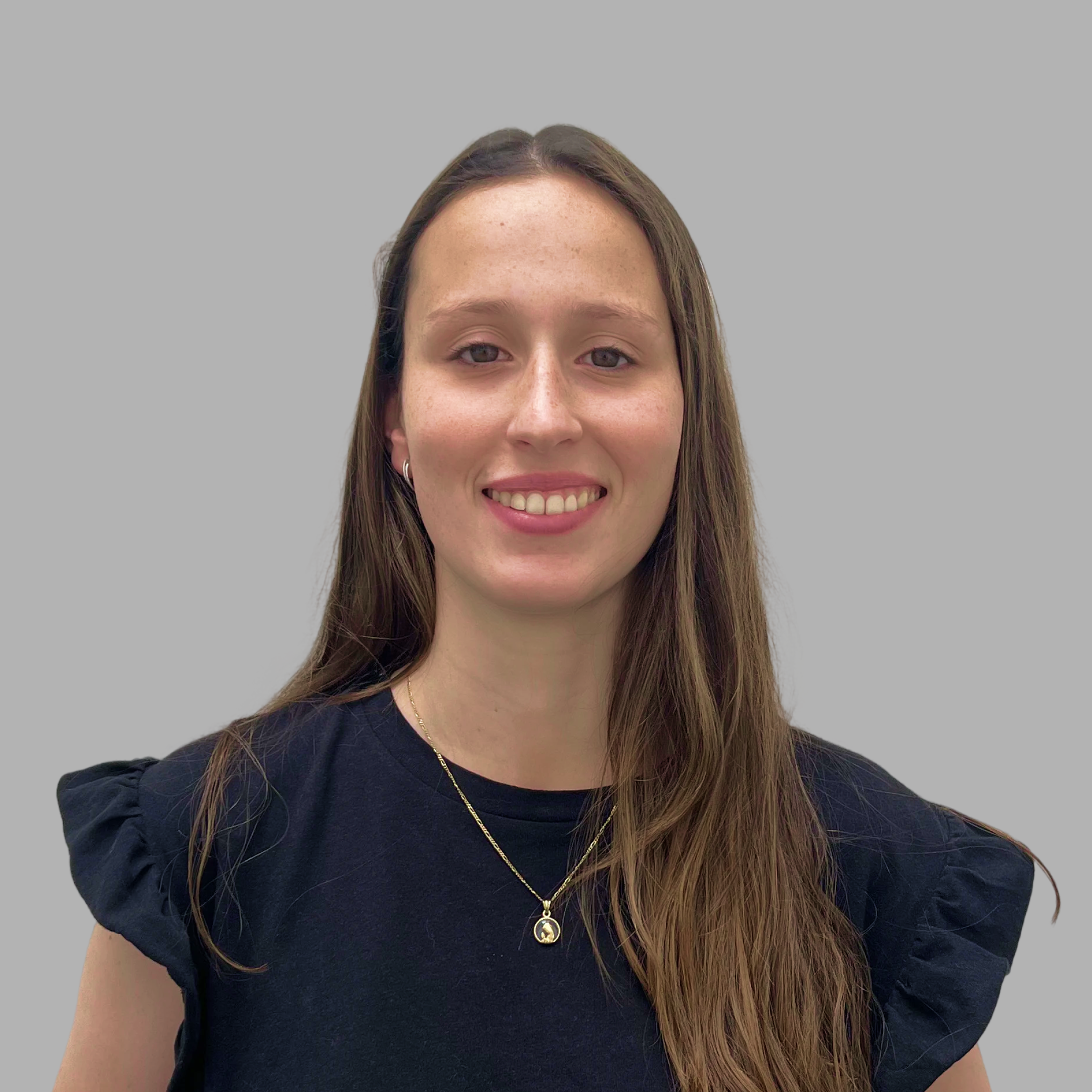 Smiling young woman with long brown hair wearing a black top and gold necklace against a gray background.