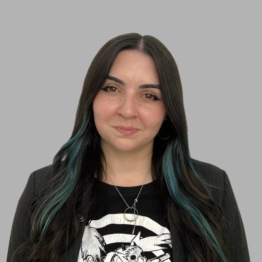 Woman with long dark hair with blue highlights wearing a black blazer and graphic T-shirt, smiling against a grey background.