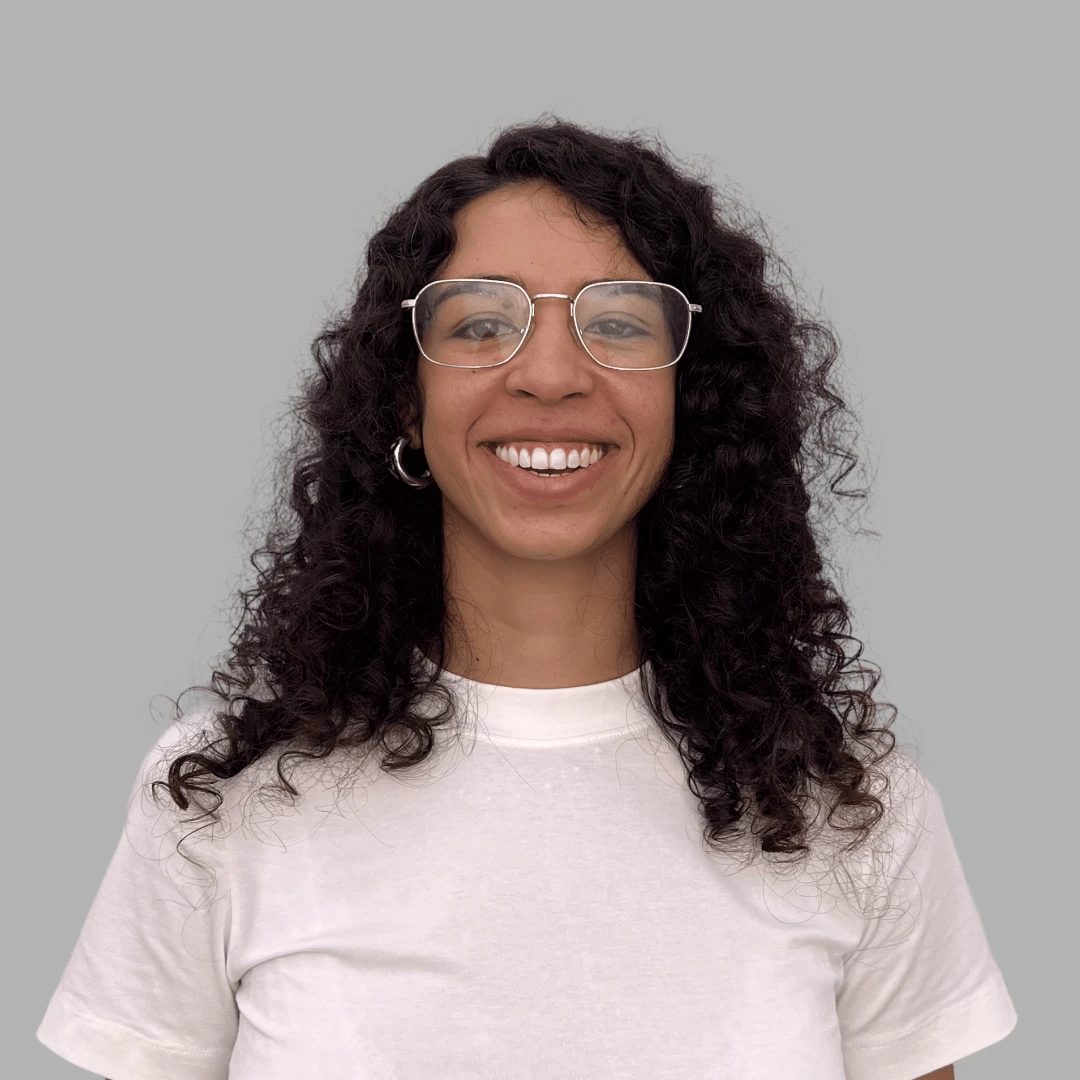 Smiling woman with curly dark hair, wearing glasses, a white t-shirt, and hoop earrings against a gray background.