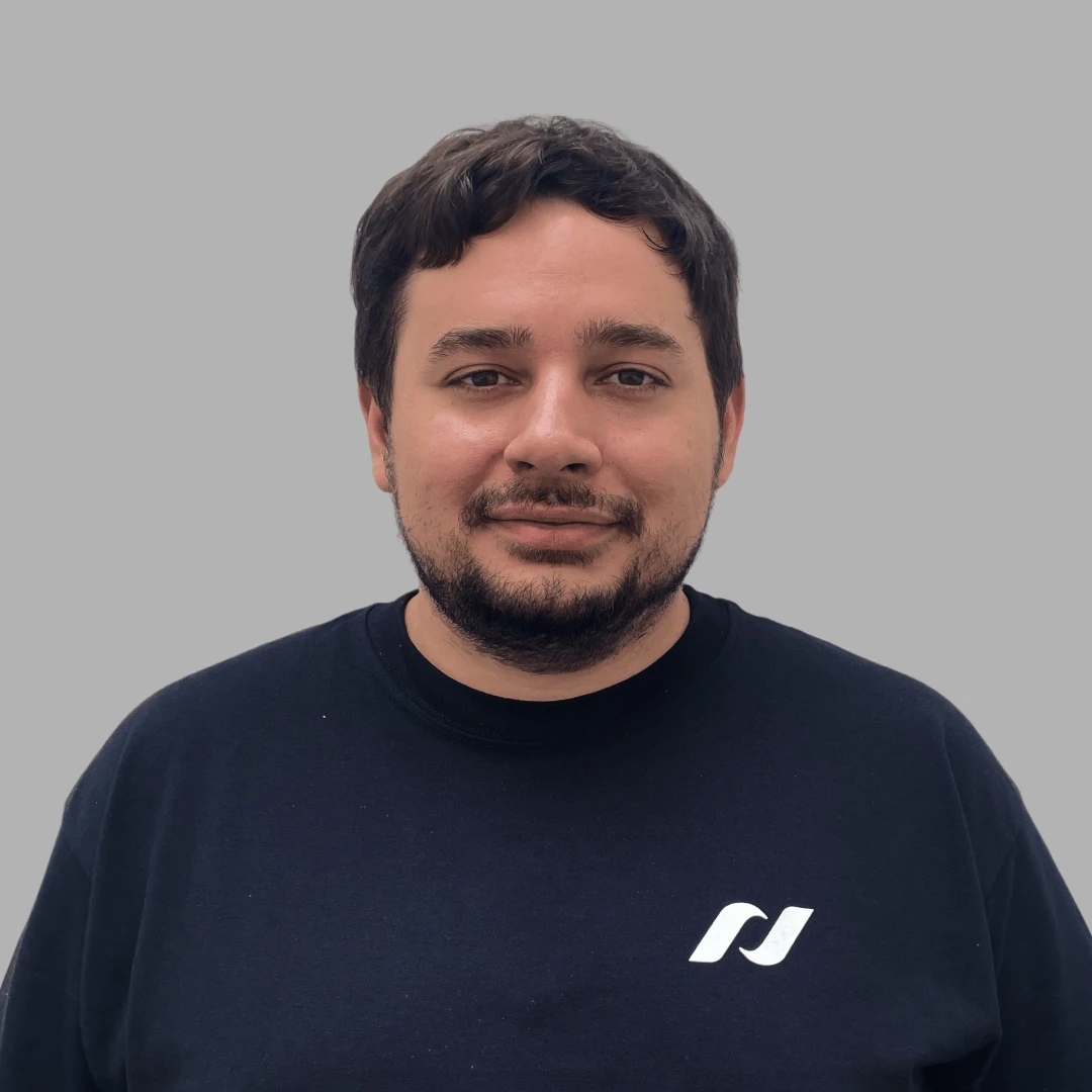 Smiling man with short dark hair and beard wearing a dark shirt with a white logo on a gray background.