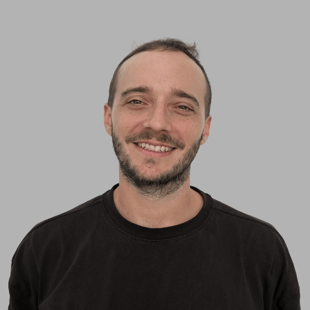 Smiling man with short hair and beard wearing a black shirt with a small red and white logo on a gray background.