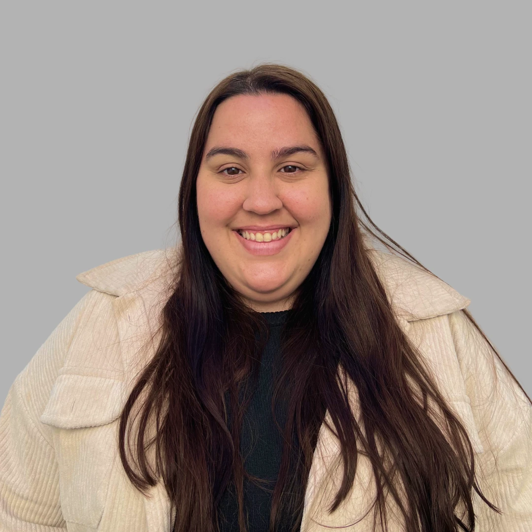 Smiling woman with long dark hair wearing a black top and light-colored jacket against a gray background.