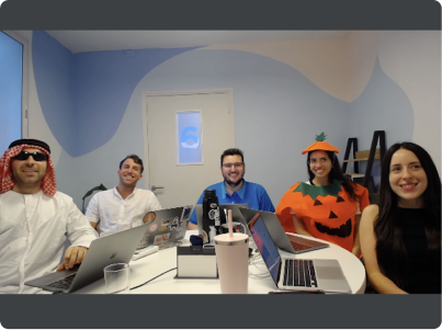 Five people sitting at a round table with laptops; one person is dressed in a pumpkin costume.