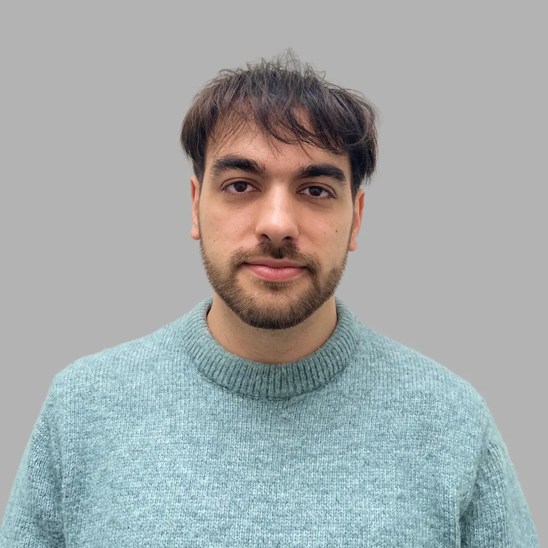 Young man with short dark hair and trimmed beard wearing a light blue knit sweater on gray background.