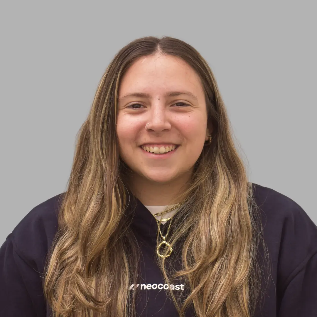 Smiling woman with long light brown hair wearing a dark shirt with the word 'neocoast' on it and gold necklaces against a gray background.