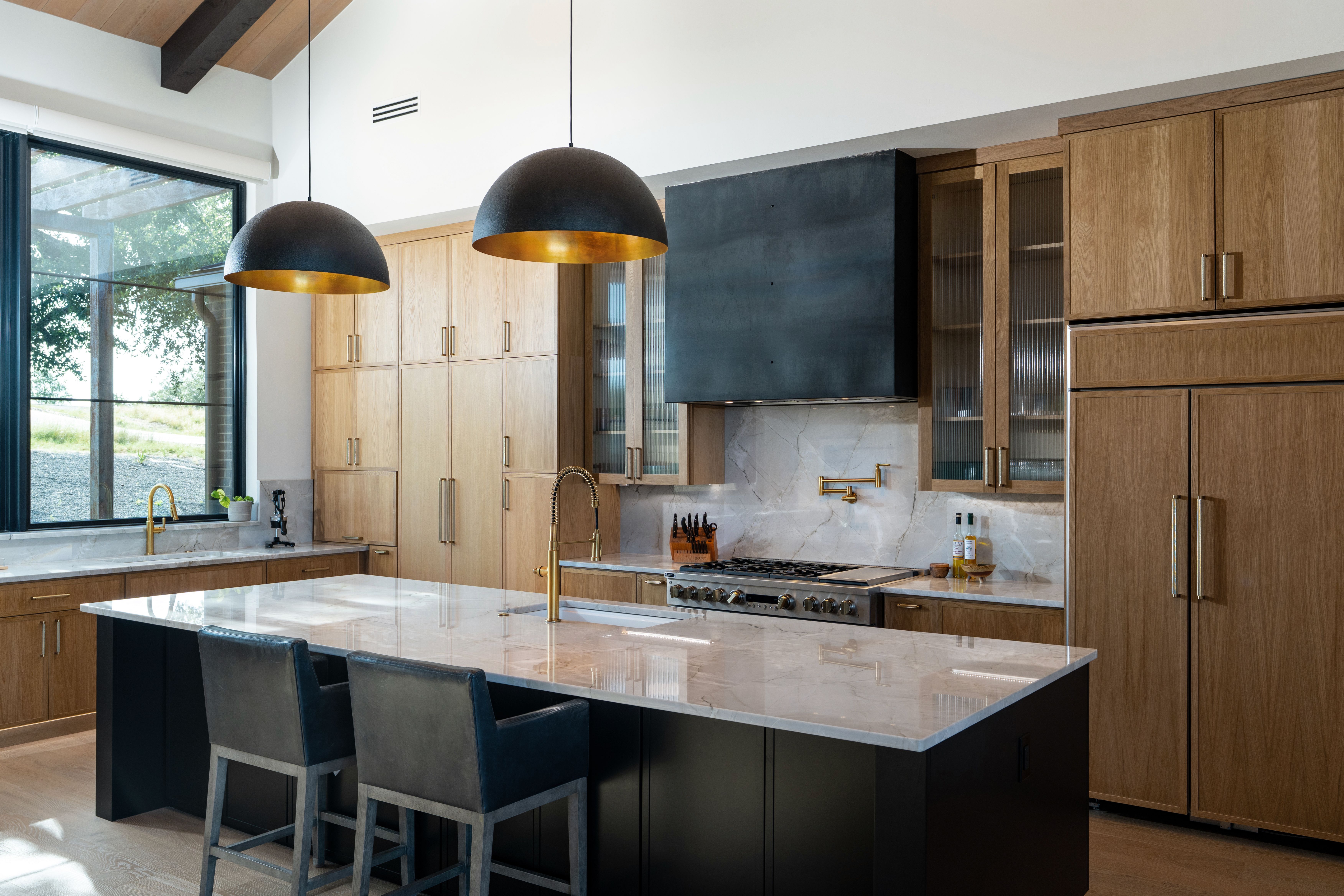 Modern kitchen with large marble island, black and gold pendant lights, wooden cabinetry, and stainless steel stove.