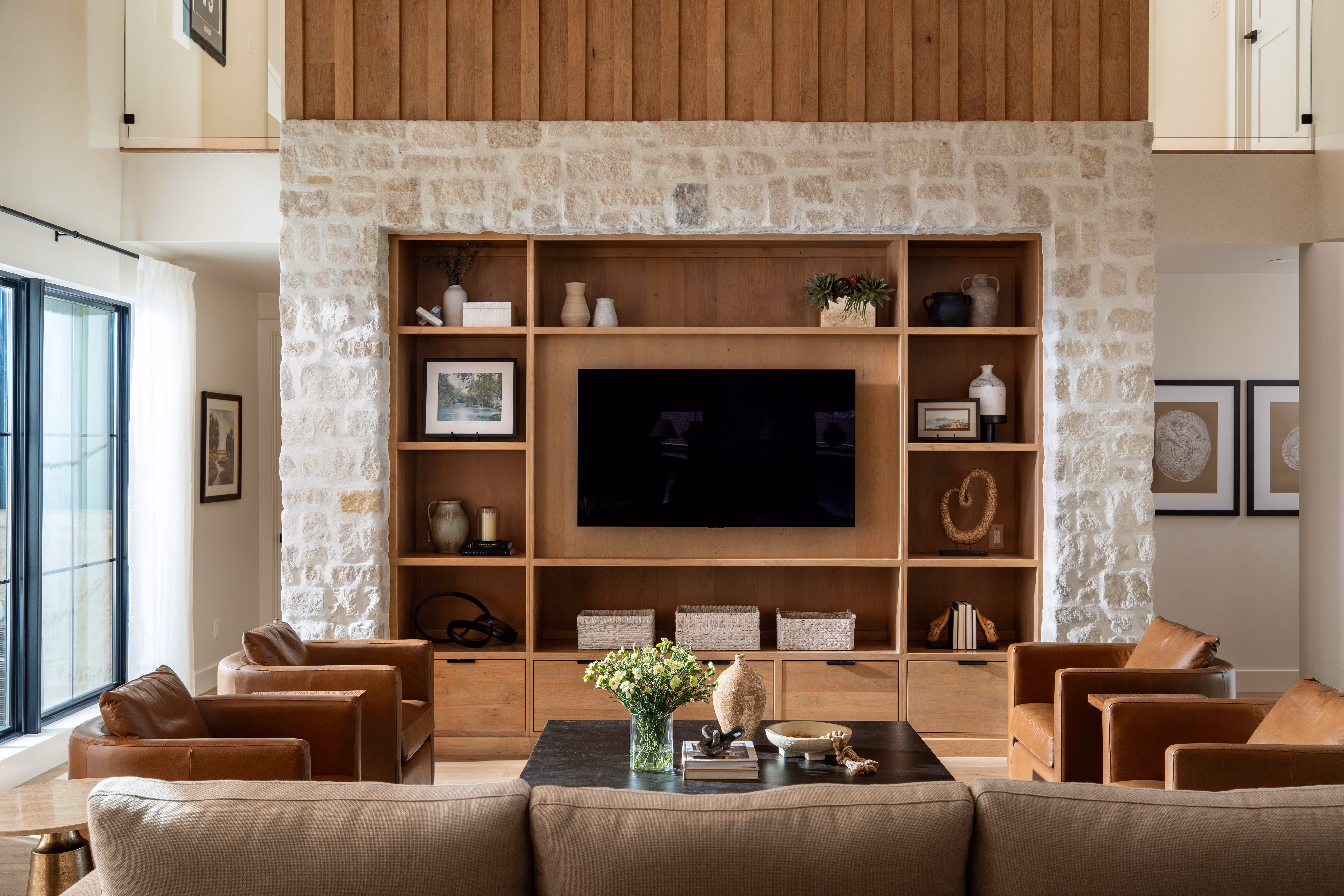 Modern living room with a beige sofa, four brown leather armchairs, a black coffee table decorated with flowers and pottery, and a wooden wall unit with a mounted TV and decorative items.