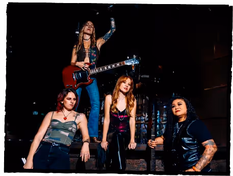 Four women posing confidently in urban setting, one holding a red electric guitar.