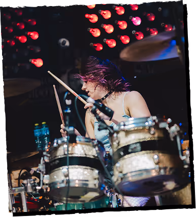 Female drummer with purple hair playing drum set on stage with red lights in the background.