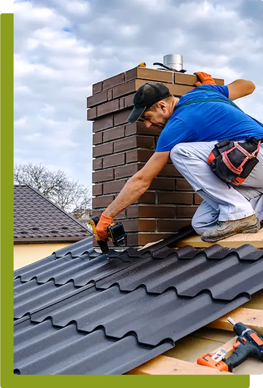 A construction worker installing metal roofing sheets around a brick chimney using a power drill. The worker is wearing gloves, a blue shirt, and a tool belt, and is kneeling on a sloped roof