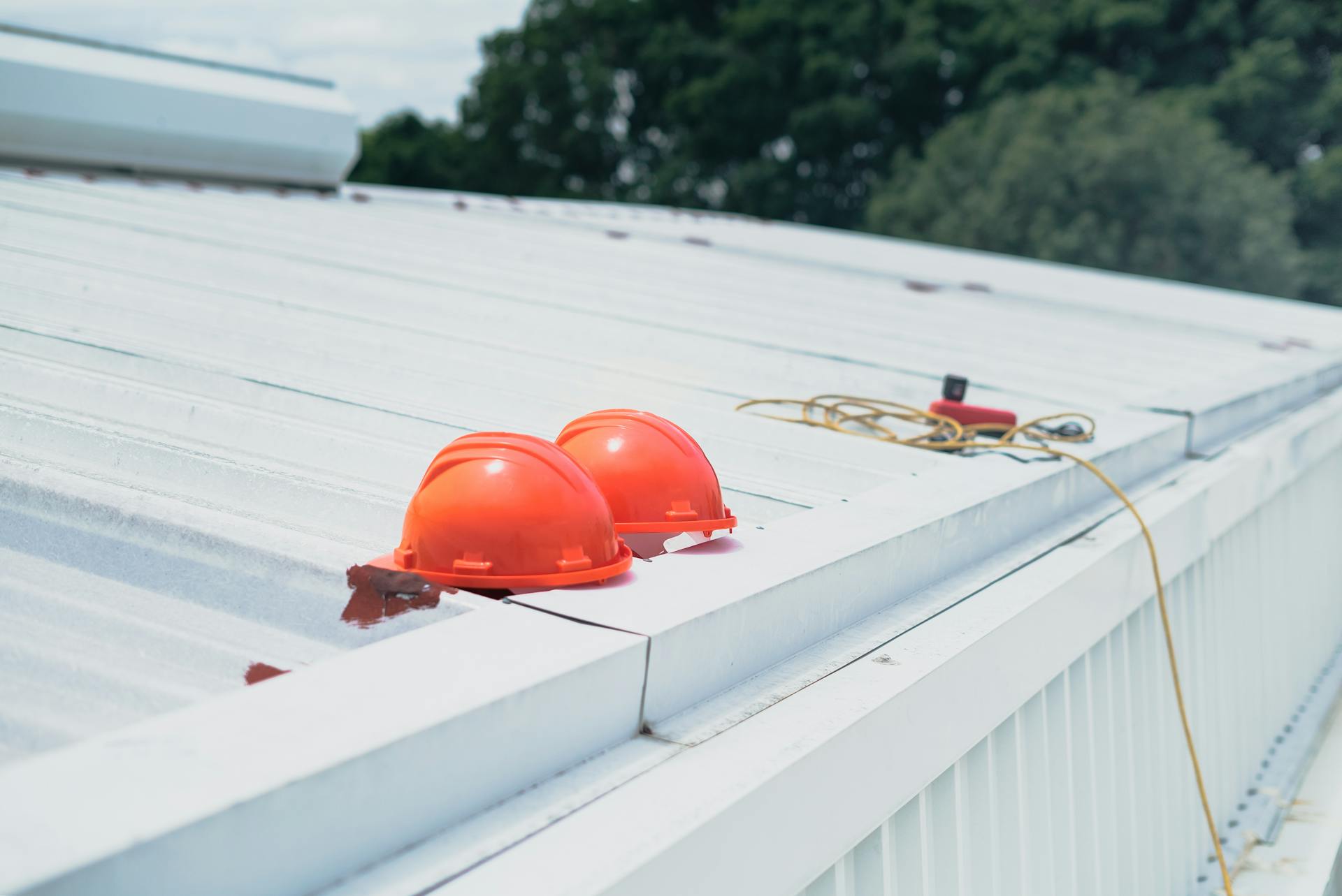 Two orange safety helmets placed on a white metal rooftop, with an electrical cord and a red device lying further along the roof. Trees are visible in the background under a partly cloudy sky.