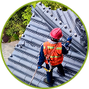 A worker wearing a red safety helmet, orange vest, and safety harness stands on a steep, dark-tiled roof, inspecting or maintaining the surface with a handheld tool. Lush green trees are visible in the background.